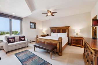 A luxurious king bedroom features a large wooden bed, elegant lighting, and a cozy chaise lounge with mountain views.