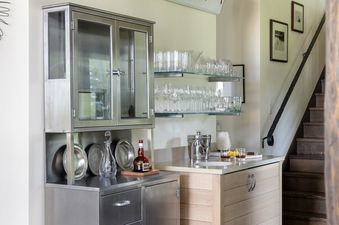 A modern bar area featuring stainless steel cabinetry, a light wood island with a sink, and glass shelves filled with glassware under a 'Please Serve Yourself' sign.