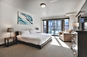 A king-sized bed with white bedding, two bedside tables with lamps, and an abstract artwork, bathed in natural light from the balcony door.