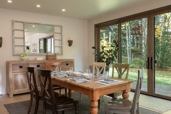 A rustic dining room features a wooden table set for a meal, a distressed wooden buffet, and large glass doors opening to a lush garden.