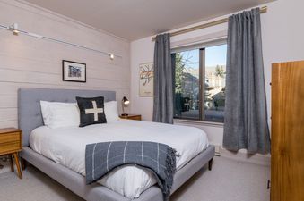 A bedroom with a large bed, a nightstand with a lamp, a dresser, and a window with a view of the mountains