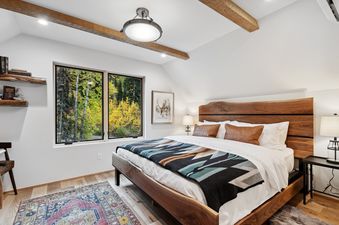 A cozy bedroom at Snowghost Chalet featuring a large wooden bed with a colorful blanket, two windows overlooking a vibrant autumn forest, and rustic wooden shelves.