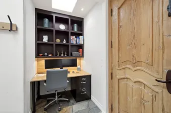 A dedicated home office at the Wintercrown Penthouse features a wooden desk with two computer monitors, a grey ergonomic chair, and a dark wood shelving unit filled with decorative items and books, alongside a rustic wooden door.