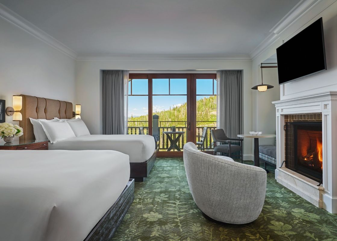 Two queen beds with white linens and brown headboards face a fireplace with a mounted television above it, and a glass door leads to a balcony with a mountain view.