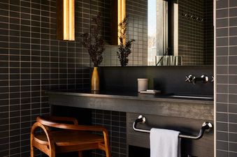 A sophisticated bathroom features dark tiled walls, a modern vanity with a large mirror reflecting a window, and warm sconce lighting.