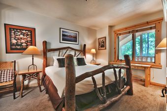 A rustic bedroom featuring a bed with a natural wood frame, white bedding, and dark pillows, flanked by wooden side tables with lamps and artwork on the walls, with a large window offering an exterior view of trees.