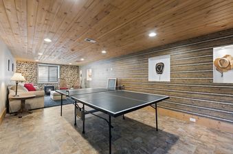 A cozy, rustic-themed lower-level living space featuring a ping-pong table, comfortable seating, a fireplace, and decorative log walls.