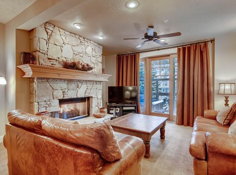 The Town Pointe B203 living room features a stone fireplace, leather sofas, a wooden coffee table, and glass doors leading to an outdoor area.