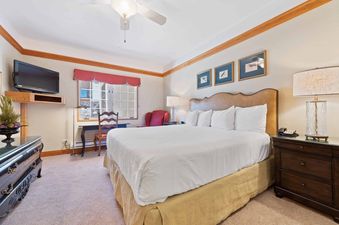 A cozy bedroom in The Charter F3105 features a king-size bed, a dark wood dresser with a TV, a desk by the window, and a ceiling fan.
