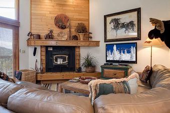 A cozy living room in Ranch at Steamboat RA510 features a brown leather sectional, a rustic wooden fireplace with a television, and a large window with outdoor views.