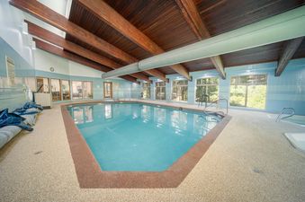 An expansive indoor pool at Beaver Village 823 features a large swimming area with clear blue water, surrounded by light blue tiled walls and a high wooden beam ceiling, with natural light filtering through multiple windows offering views of lush greenery