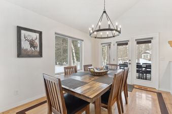 A rustic wooden dining table surrounded by chairs under a stylish chandelier, with large windows overlooking a snowy winter landscape.
