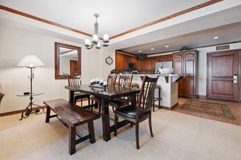 A cozy dining area features a dark wood table and bench, with a full kitchen visible in the background, all bathed in soft lighting.