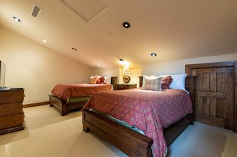 Two large beds with red and gold patterned blankets and throw pillows, with a wooden headboard and base, in a room with a light cream sloped ceiling and light cream walls.