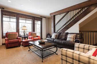A cozy living room at Silver Star 902 features dark wood accents, a brown leather sofa, red leather armchairs, and a wrought-iron coffee table, with large windows overlooking a snowy landscape.