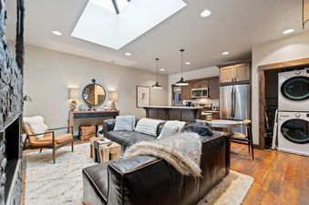 A bright and airy open-concept living area in Central Loft 303 featuring a dark leather sofa, a cozy armchair, a modern kitchen with wooden cabinetry, and a stacked stone fireplace.