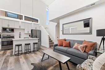 A modern open-concept living and kitchen area featuring white cabinetry, stainless steel appliances, a gray sofa with orange pillows, a coffee table, and a cowhide rug.
