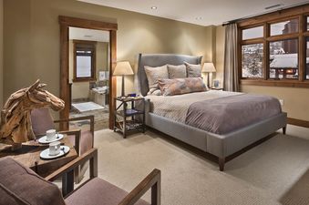A cozy bedroom is elegantly furnished with a large bed, two armchairs, a small table, and a unique wooden horse sculpture, offering a view into an adjacent bathroom and a snowy outdoor landscape through multiple windows.