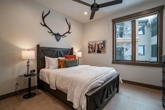 A cozy bedroom in Elevated Vibes featuring a dark wooden bed, wooden antler decor, and modern paintings, with a window offering a view of nearby buildings and snow-covered eaves.