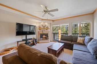 A cozy living room at St James Place 325 features a fireplace, a flat-screen TV, and comfortable sofas, with a large window offering a view of lush trees outside.