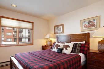 A rustic bedroom at Ranch at Steamboat RA604 with a red and black plaid bed, wooden furniture, and a window overlooking a snowy exterior.