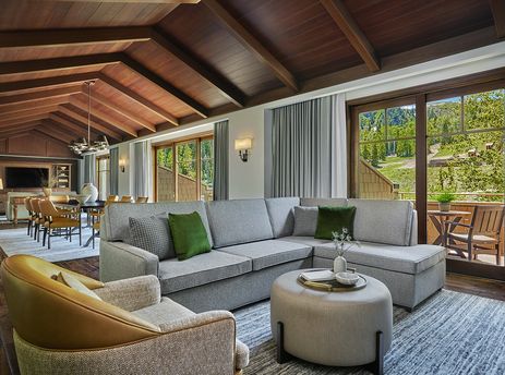 A spacious and inviting living area in the Montage Flagstaff Suite features a comfortable sectional sofa, a round ottoman, and a dining area, all under a vaulted wooden ceiling, with large windows overlooking a lush green mountain landscape.