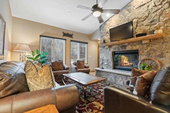 A cozy living room at The Charter B4305 with a stone fireplace, television, leather sofas, armchairs, and large windows looking out to a winter landscape.
