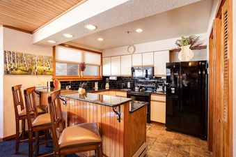 The kitchen at Chateau Chamonix CX134 features black appliances, wooden cabinets, a black subway tile backsplash, and a breakfast bar with bar stools, all under a distinctive wooden plank ceiling.