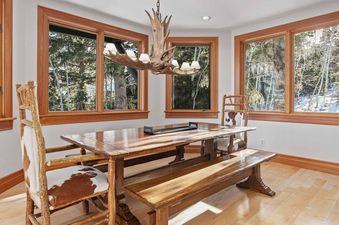 The dining area at 7780 Sterling Drive features a large wooden table, two chairs with cowhide upholstery, a wooden bench, and a unique antler chandelier, all set against a backdrop of windows showcasing snow-covered trees.