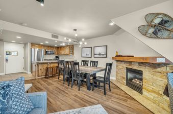 An inviting open-concept living space in Lowell Slopeside 260, featuring a dining area, a fully equipped kitchen, and a stone fireplace, all with rustic and modern furnishings.