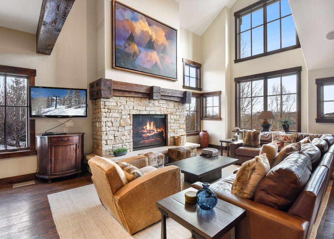 A spacious and luxurious living room at Silver Star 47 featuring a stone fireplace, high ceilings, large windows with winter views, and comfortable leather seating.