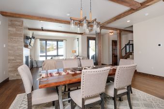 A rustic dining area with a wooden table and upholstered chairs, leading to a living room with mountain views, within Mountain Lake Condo 101B.