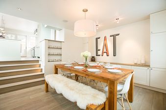 A dining room with a large wooden table and white chairs, a white fur bench, and white cabinets