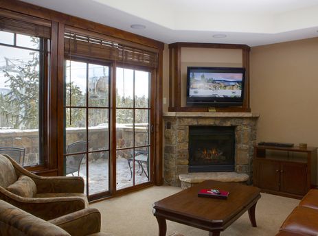 A cozy living room in the Crystal Peak 3 Bedroom unit, featuring a stone fireplace, a flat-screen TV, comfortable sofas, and large windows looking out onto a snow-covered landscape with a gondola.