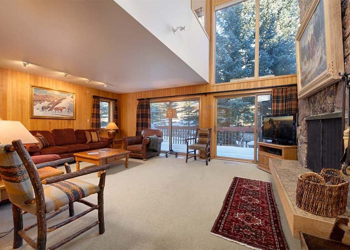 Comfortable living room at Windriver 1 with a stone fireplace, wood paneling, and views of snowy trees through large windows.