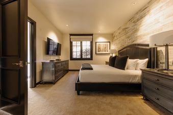 A luxurious bedroom at Red Hawk Retreat at See Forever features a bed with a black headboard, a dark dresser with a TV, a window, and a rustic wooden accent wall.