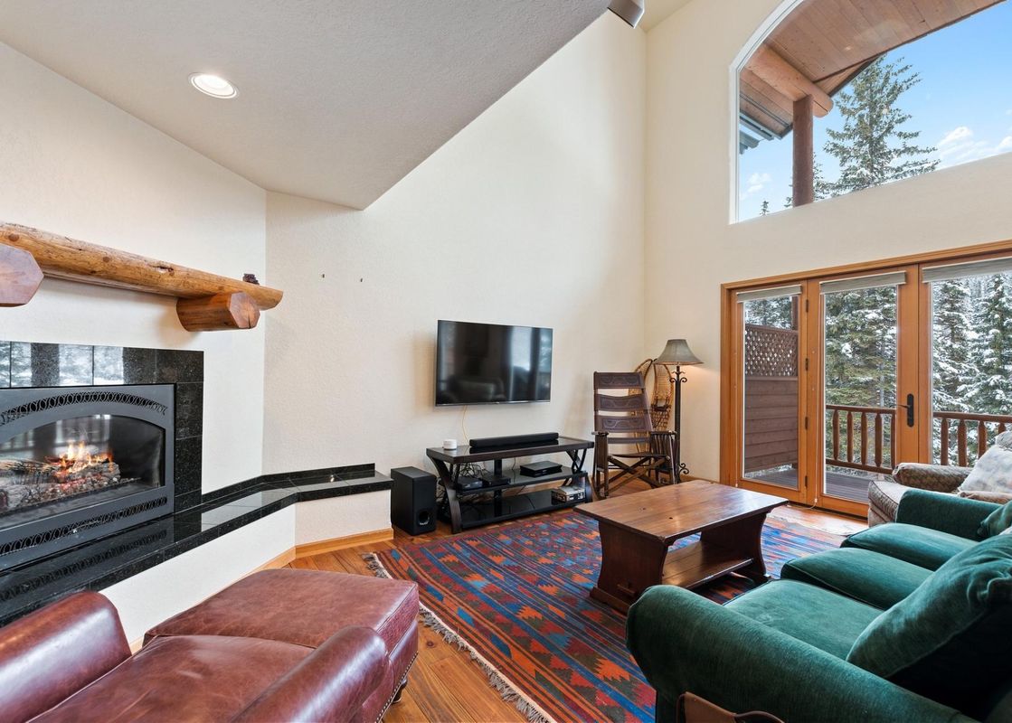 The living area at Silvertip Ski Haus features a fireplace, TV, comfortable seating, and a view of snowy pine trees through a large window.