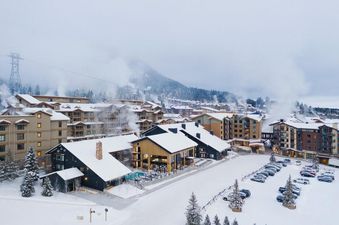 Winter aerial view of the snow-covered Gravity Haus JH Two Queens resort complex with mountains in the background.