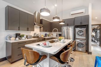 A modern kitchen with stainless steel appliances, a white quartz island with bar stools, and a washer and dryer combo stacked on top of each other.