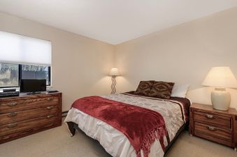 A cozy bedroom with a bed featuring a red throw, two nightstands with lamps, a dresser with a television, and a window.