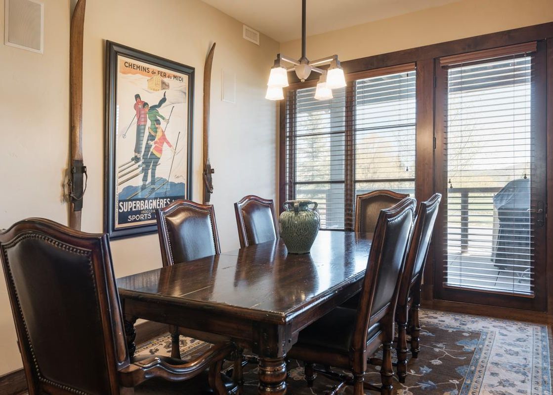 A rustic dining area with a large wooden table, surrounded by leather chairs, under a hanging chandelier, with a ski-themed poster on the wall.