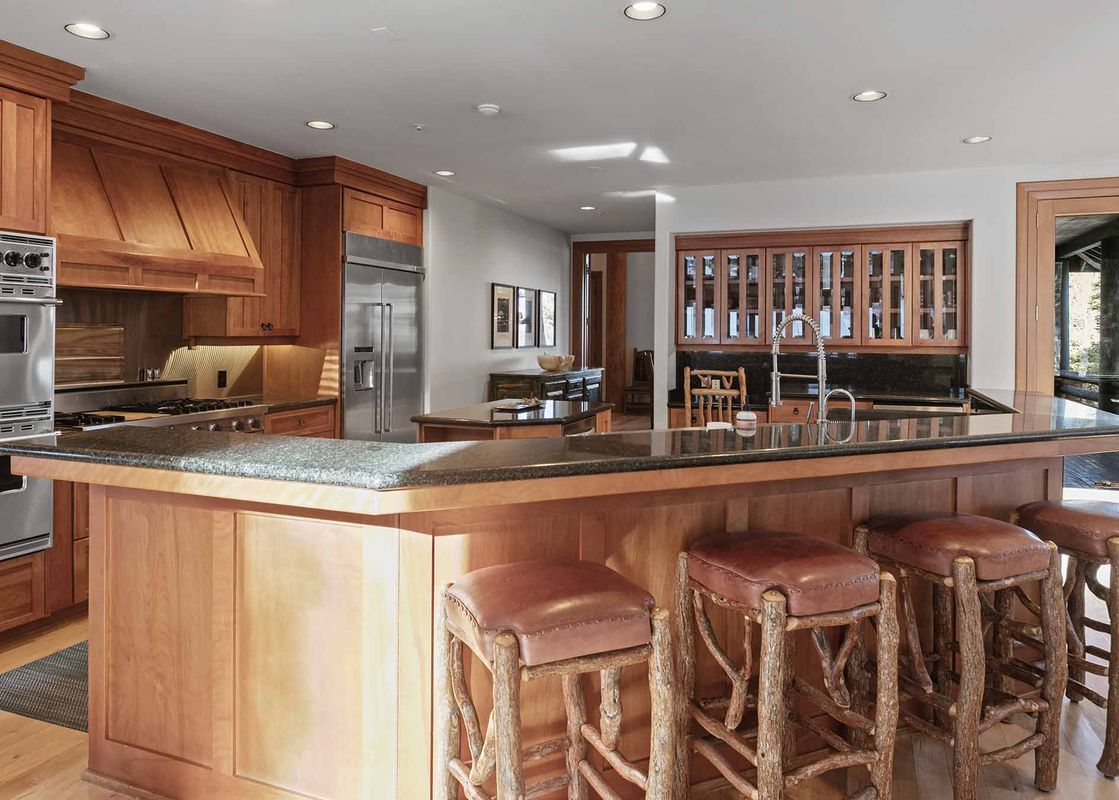 A luxury kitchen with warm wood cabinetry, stainless steel appliances, a large granite island with a sink, and three rustic wooden bar stools with leather seats.