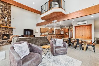 A spacious, open-concept living area with a stone fireplace, comfortable seating, a dining table, and a modern kitchen, all under a mezzanine with a black railing, at the Chair View Chalet.