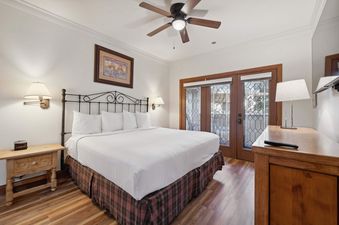 A cozy bedroom with a large bed, decorative headboard, wooden nightstands, and a ceiling fan, featuring double glass doors leading to a balcony.