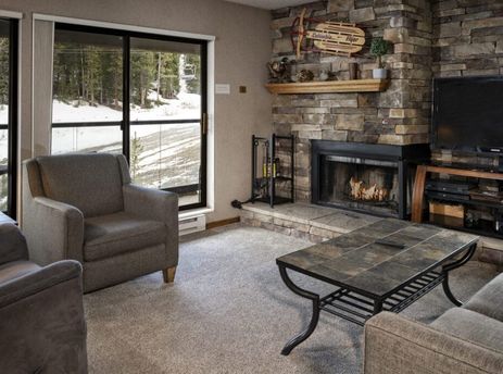 A cozy living room featuring a stone fireplace, comfortable seating, and large windows with a view of a snowy, tree-lined landscape.