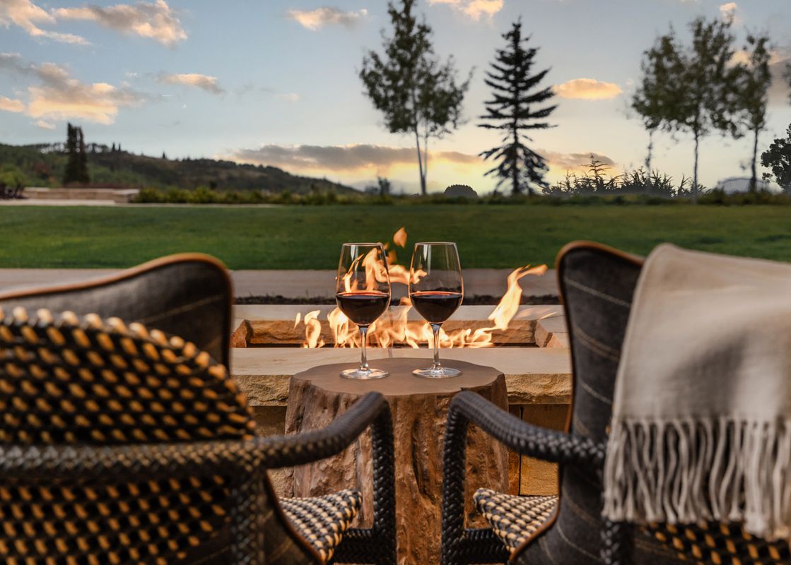 Two wine glasses with red wine rest on a rustic wooden table in front of a flickering fire pit, flanked by two comfortable chairs, with a lush green lawn and distant trees under a soft, cloudy sky at sunset.
