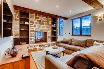The living area features a stone fireplace, comfortable L-shaped sofa, and large windows with mountain views.