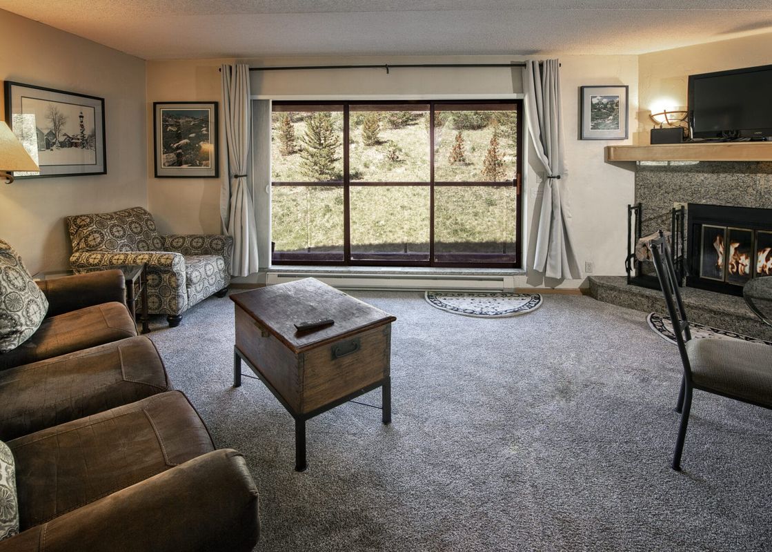 A comfortable living room in the Colorado Suite Beaver Run Resort features a brown sofa, a patterned armchair, a wooden coffee table, and a granite fireplace with a mounted TV, all overlooking a scenic outdoor landscape through large windows.
