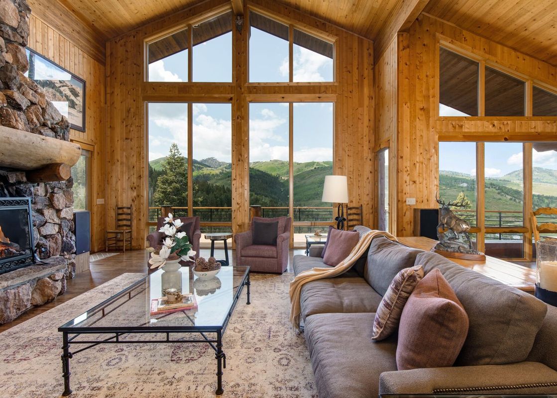 A spacious and inviting lounge at American Flag Solamere, featuring a large L-shaped sofa, a glass coffee table, two armchairs, a stone fireplace, and panoramic mountain views through floor-to-ceiling windows.