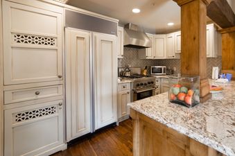 A fully equipped kitchen features custom cream cabinetry, stainless steel appliances including a large refrigerator and a range with an overhead hood, light granite countertops, and wood flooring, all illuminated by recessed ceiling lights.